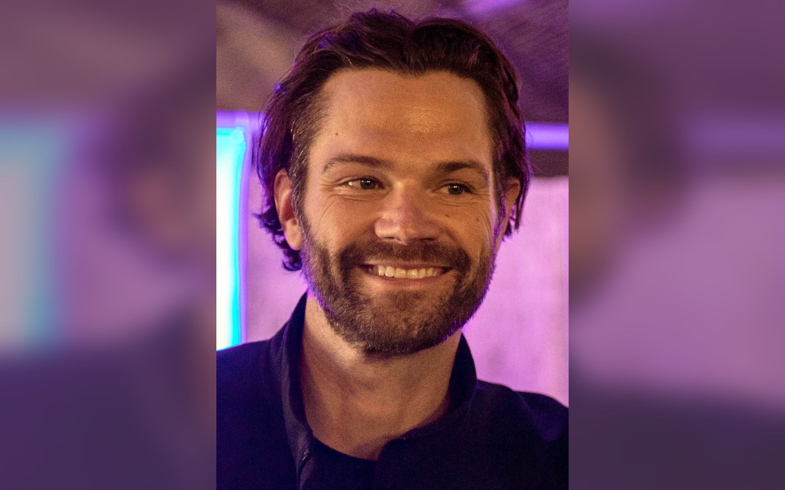 Jared Padalecki smiling with a beard, lit by purple ambient lighting
