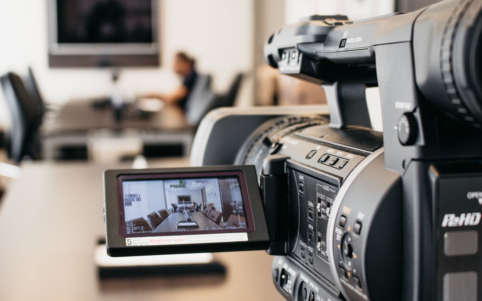 A video camera with flip-out screen recording a meeting room with a person seated at a table