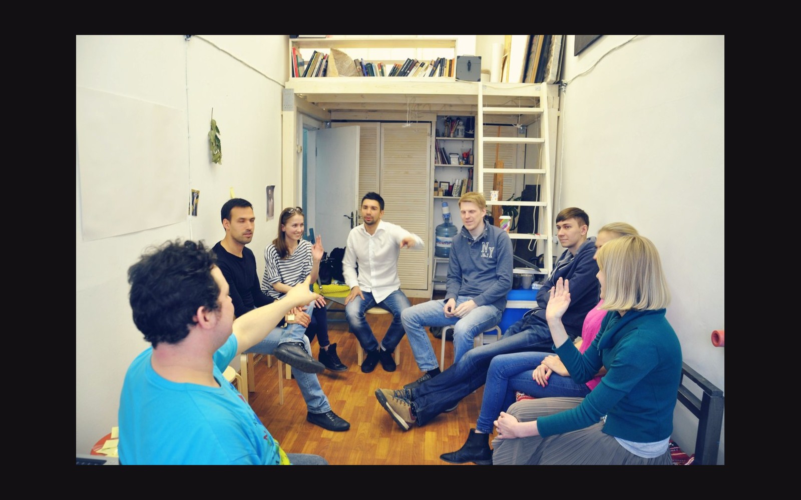 A group of young adults sit in a circle discussing in a small studio room